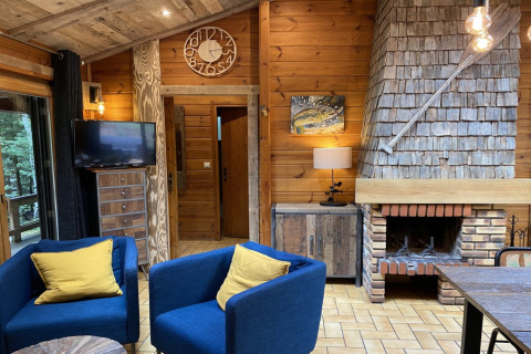 Woonkamer in het LE FISHING LODGE houten chalet in Frankrijk met open haard, blauwe stoelen en gele kussens.