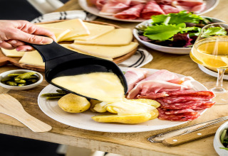 Gesmolten kaas wordt over aardappelen gegoten, geserveerd met vleeswaren, salade en witte wijn in een chalet.