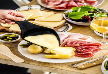 Gesmolten kaas wordt over aardappelen gegoten, geserveerd met vleeswaren, salade en witte wijn in een chalet.