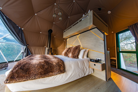 Modern bedroom with plush bedding, wood stove, and geometric windows in Chartwell Luxury Dome, UK.
