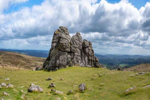 Large rocky outcrop standing on a grassy plain with dramatic clouds above and rolling hills in the distance.