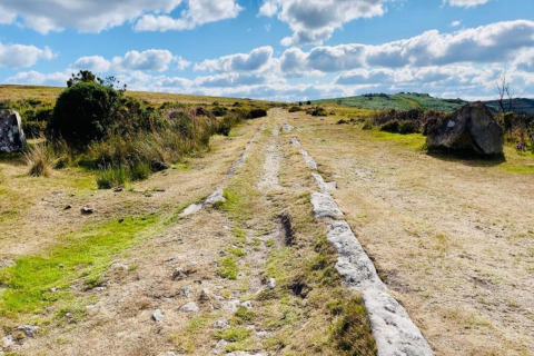 Ancient stone tracks cut through open grassland under a bright blue sky with clouds and distant hills.