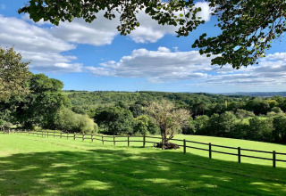 Zicht vanaf een koepel op groene weiden, bomen, een houten omheining en blauwe hemel met wolken.