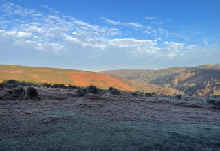 Scenic rolling hills with green and reddish tones beneath a blue sky dotted with fluffy white clouds.