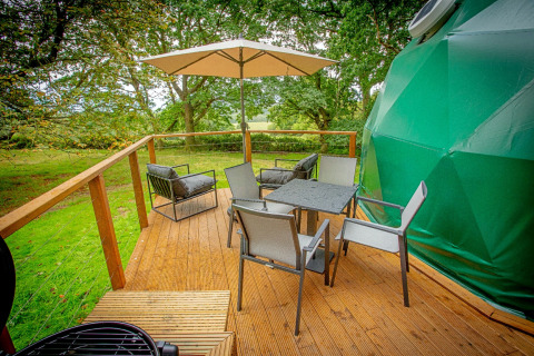 Udendørs terrasse ved Hever Luxury Dome i Storbritannien med stole, parasol og udsigt til træer.