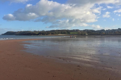 Photo of an open sandy beach with shallow water, a few distant people, and a cloudy blue sky above.