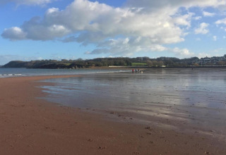 Photo of an open sandy beach with shallow water, a few distant people, and a cloudy blue sky above.