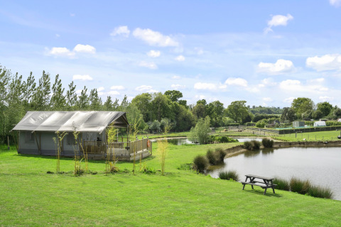 Het Herdwick Safari Tent naast een vijver met picknicktafel, op een groene weide in het Engelse platteland.