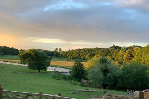 Uitzicht op een groen veld, rivier en bos bij zonsondergang vanaf een glamping accommodatie.