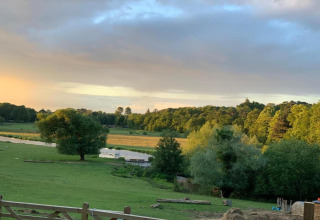 Uitzicht op een groen veld, rivier en bos bij zonsondergang vanaf een glamping accommodatie.