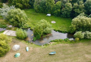 Luchtfoto van een glampinglocatie met tenten, bootjes en een caravan bij een rivier omringd door bomen.
