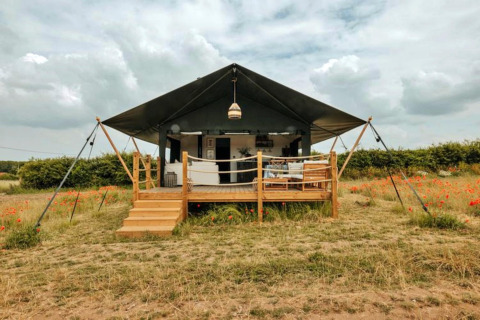 Safaritent Owls Nest met houten veranda, gelegen in een veld met wilde bloemen onder een bewolkte lucht.