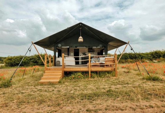 Safaritent Owls Nest met houten veranda, gelegen in een veld met wilde bloemen onder een bewolkte lucht.