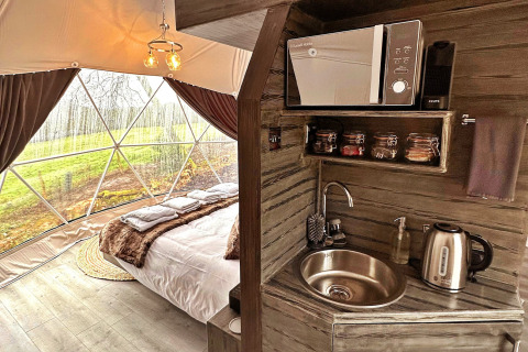 Interior view of Ashdown Luxury Dome in the UK, showing a cozy bed and small kitchenette with amenities.