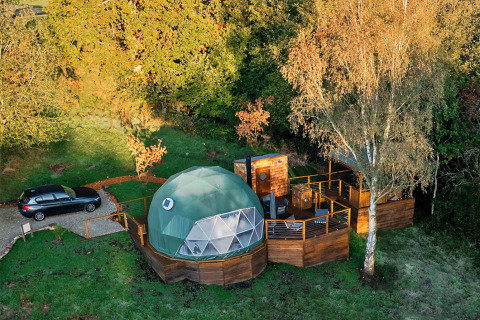 Aerial view of Wakehurst Luxury Dome, a green geodesic dome with wooden deck and car, surrounded by trees.