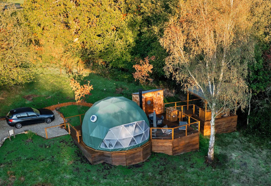 Luchtfoto van de Wakehurst Luxury Dome, groene koepel met houten terras en auto, omringd door bomen.