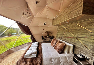Interior of Wakehurst Luxury Dome in the UK with a cozy bed, stylish decor, and panoramic green views outside.