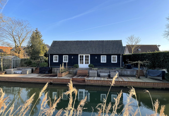 Udendørsbillede af ferieboligen De Vlonder med træterrasse ved vandet på en klar dag.