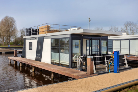 Moderne husbåd ved kaj, med store vinduer og terrasse, fortøjet på roligt vand i Holland.