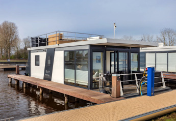 Moderne husbåd ved kaj, med store vinduer og terrasse, fortøjet på roligt vand i Holland.