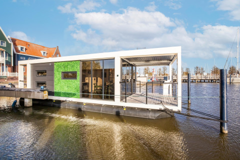 Moderne husbåd ved en mole, med grøn væg og store vinduer, fortøjet ved marinaen under blå himmel.