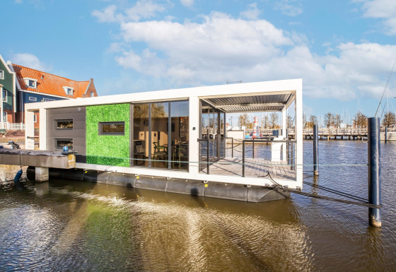 Moderne husbåd ved en mole, med grøn væg og store vinduer, fortøjet ved marinaen under blå himmel.