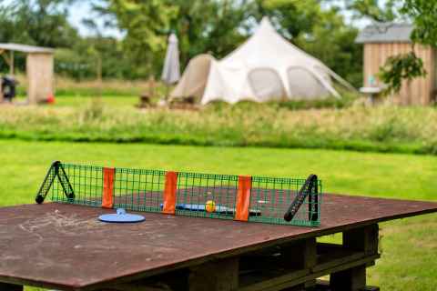 Set da ping pong su tavolo di legno all’aperto, con grande tenda teepee sul prato sullo sfondo.