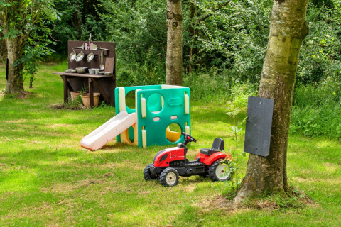 Area giochi per bambini in giardino con scivolo, cucina giocattolo e trattore rosso tra gli alberi.
