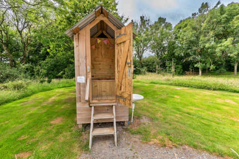Bagno esterno in legno presso un campeggio con tenda teepee, circondato da prati e alberi verdi.