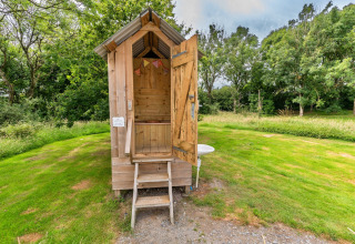 Bagno esterno in legno presso un campeggio con tenda teepee, circondato da prati e alberi verdi.