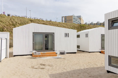 Moderne strandhuse i hvide træpaneler på sandet ved Beach Houses Zandvoort 2 med hotel i baggrunden.