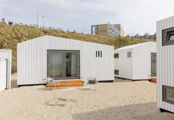Moderne strandhuse i hvide træpaneler på sandet ved Beach Houses Zandvoort 2 med hotel i baggrunden.