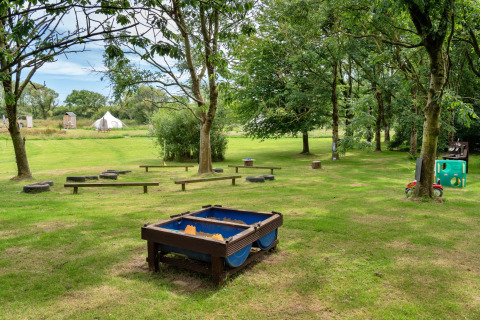 Area giochi con sabbiera, panche di legno e pneumatici, circondata da alberi e una tenda tipi sullo sfondo.