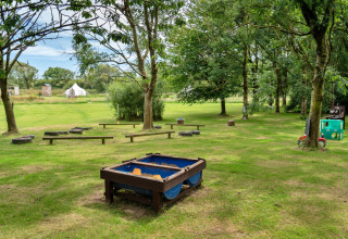 Area giochi con sabbiera, panche di legno e pneumatici, circondata da alberi e una tenda tipi sullo sfondo.