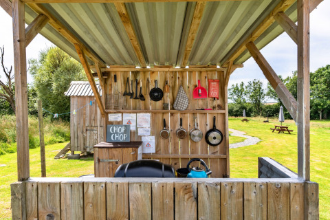 Cucina all'aperto accanto a una tenda tipi con pentole, utensili e barbecue sotto una tettoia su prato.