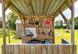 Cucina all'aperto accanto a una tenda tipi con pentole, utensili e barbecue sotto una tettoia su prato.
