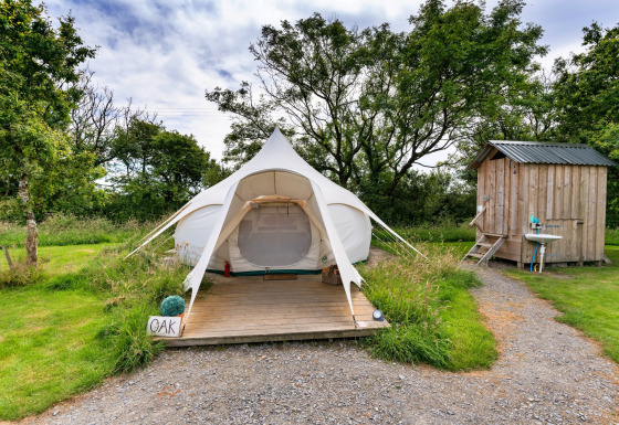 Foto di una tenda tipi Oak Lotus Belle con pedana in legno e una piccola casetta in un campeggio verde.