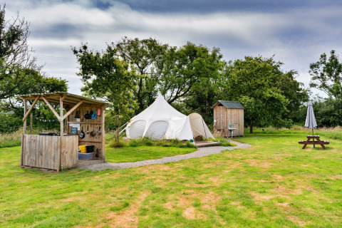 Glamping in Regno Unito con tenda Oak Lotus Belle, cucina esterna, cabina e tavolo da picnic su prato verde.