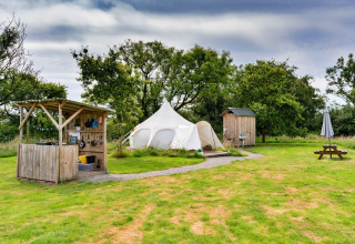 Glamping in Regno Unito con tenda Oak Lotus Belle, cucina esterna, cabina e tavolo da picnic su prato verde.