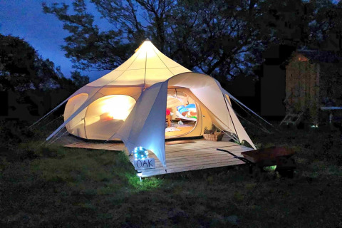 Tenda tipi Oak Lotus Belle illuminata nel Regno Unito di notte, atmosfera accogliente e alberi sullo sfondo.