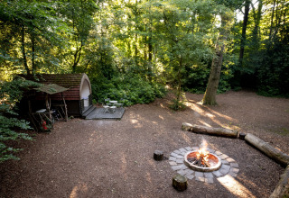 Glamping-pod in het bos met kampvuur, houten zitbanken en buitenterras in het daglicht.