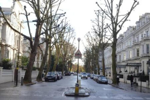 Bygade i Storbritannien med parkerede biler, hvide bygninger og løvfrie træer på begge sider.