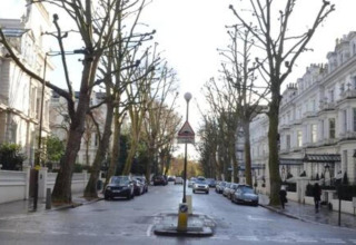 Bygade i Storbritannien med parkerede biler, hvide bygninger og løvfrie træer på begge sider.