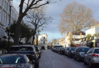 En stille britisk gade med parkerede biler på begge sider under store, bladløse træer og skyet himmel.