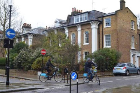 To cyklister kører forbi vejskilte foran et klassisk britisk hus i en rolig gade i Storbritannien.