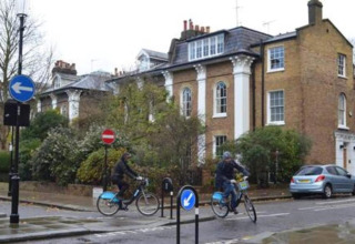 To cyklister kører forbi vejskilte foran et klassisk britisk hus i en rolig gade i Storbritannien.
