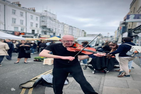 En gademusiker spiller violin midt på en travl markedsplads med folk i baggrunden i Storbritannien.
