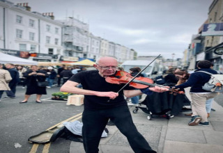 En gademusiker spiller violin midt på en travl markedsplads med folk i baggrunden i Storbritannien.