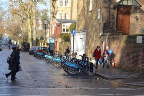 Folk går forbi en cykeludlejning på gaden i Storbritannien, med træer og bygninger omkring dem.