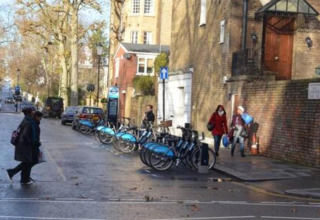 Folk går forbi en cykeludlejning på gaden i Storbritannien, med træer og bygninger omkring dem.